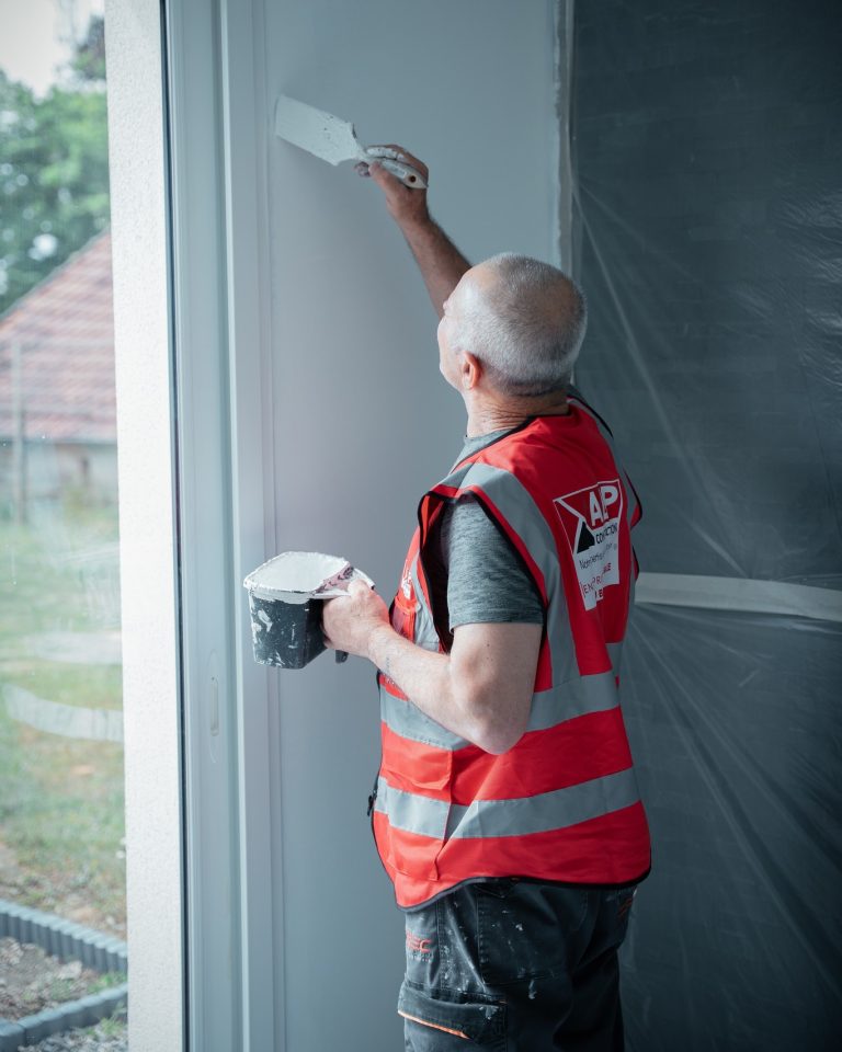 Un peintre ALP pr&eacute;pare et peint un mur int&eacute;rieur, en gilet rouge au logo ALP.