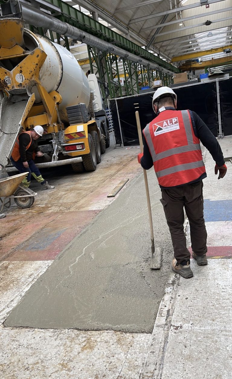 Un salari&eacute; ALP en gilet rouge et casque travaille le b&eacute;ton frais &agrave; proximit&eacute; d&rsquo;un camion-toupie.